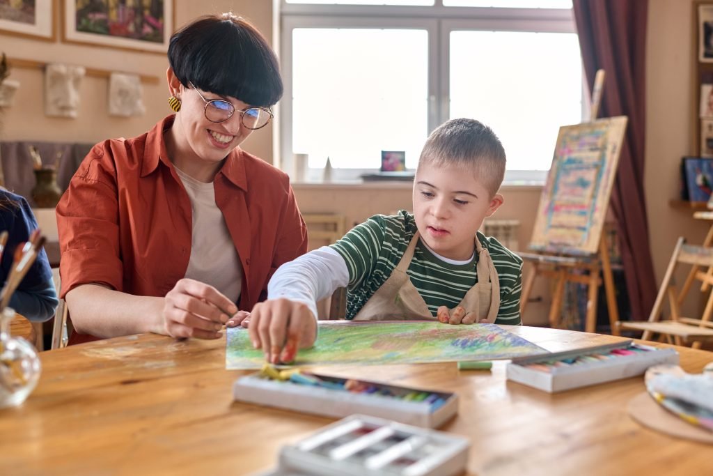 Young boy with Down syndrome enjoying an art lesson, drawing with crayons while his teacher helps