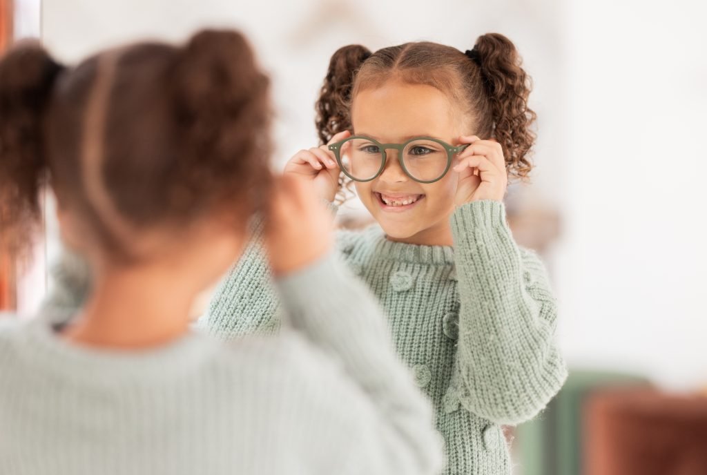 Smiling girl trying on new glasses and looking at her reflection in a mirror at an optical store