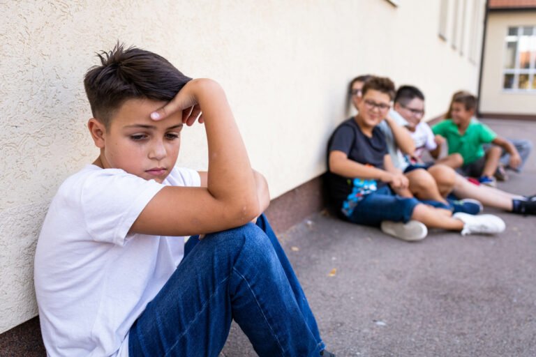 Boy sitting alone against a school wall while a group of students sits together nearby