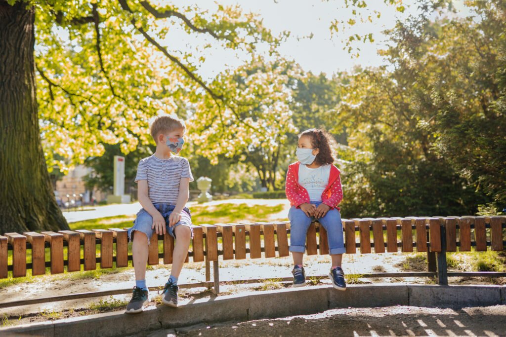 Two children in face masks sit far apart on a park bench under a leafy tree