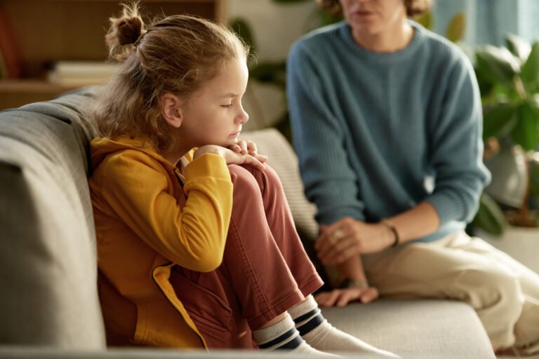 A young girl in a yellow hoodie sits curled up on a sofa while a caring adult sits close beside her